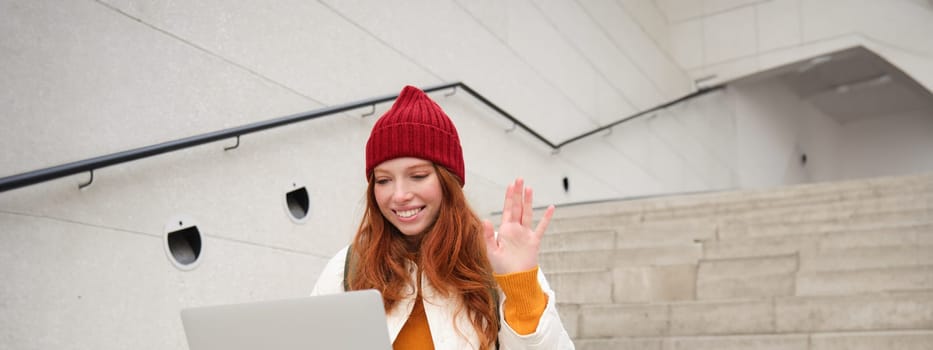 Portrait of happy young woman, redhead girl with laptop, waves hand and says hello on video chat, has conversation on computer application, talks to someone.