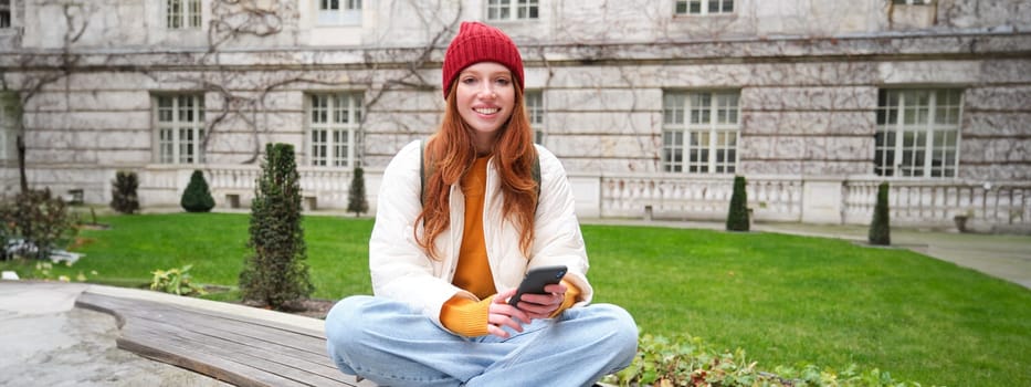 Portrait of stylish young woman, 25 years, sits on bench in park and uses mobile phone, reads online news, messages or watches video on smartphone app, connects to public wifi.