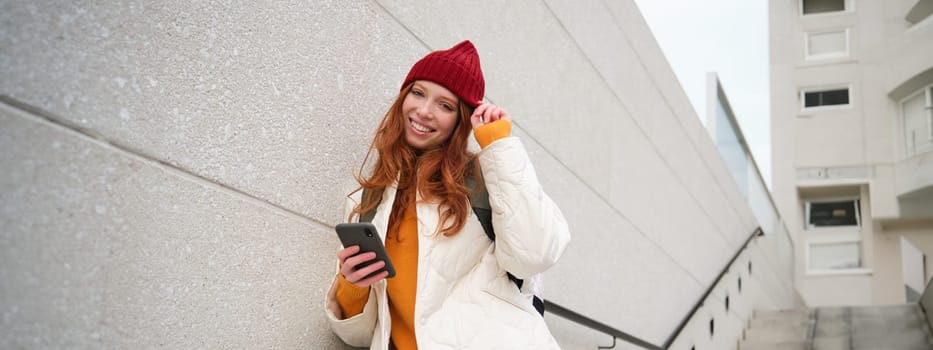 Happy girl student in red hat, holds smartphone, tourist looks at map app on her phone, explores sightseeing, texts message, looks for couchsurfing, rents place to stay online.