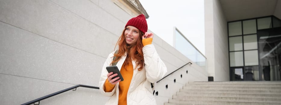 Happy girl student in red hat, holds smartphone, tourist looks at map app on her phone, explores sightseeing, texts message, looks for couchsurfing, rents place to stay online.