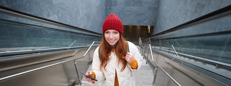 Portrait redhead girl tourist, goes up stairs with smartphone, follows route on mobile phone app, holds backpack and smiles.