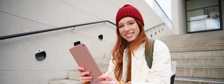 Happy stylish redhead girl, student in red hat, holds digital tablet, uses social media app, searches something online, connects to wifi.