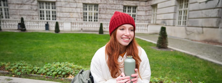 Smiling redhead girl rests in park, sits on bench with backpack, drinks from thermos, enjoys hot drink from flask and looks relaxed.