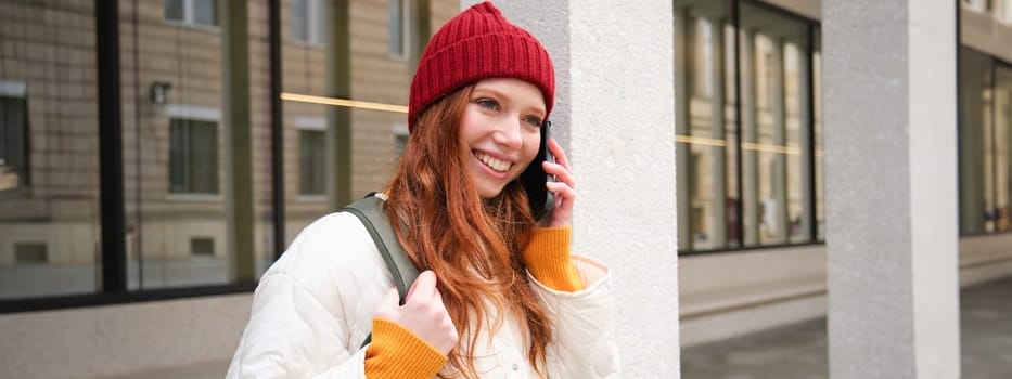 Portrait of smiling ginger girl with red hat, stands on street with backpack, rings someone on phone app, talks on mobile, uses smartphone.