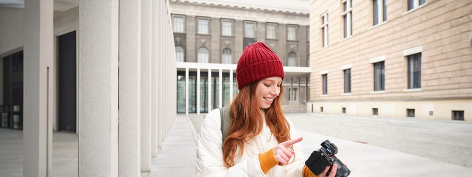 Smiling redhead girl photographer, taking pictures in city, makes photos outdoors on professional camera. Young talent and hobby concept