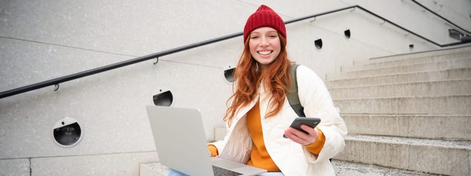 Young tourist, girl booking hotel room on laptop, enters confirmation code on smartphone, sitting with computer and phone on stairs outdoors.