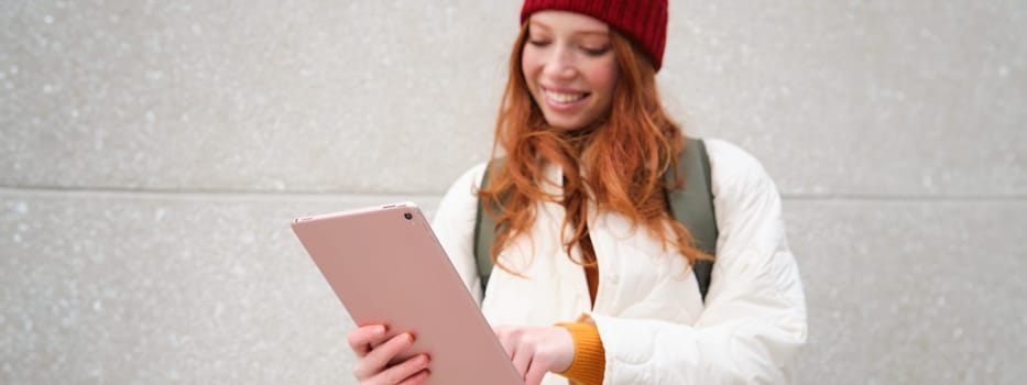 Young redhead girl, walks with digital tablet around city, stands on street with backpack and gadget, looks at online map, searches for a place in internet.