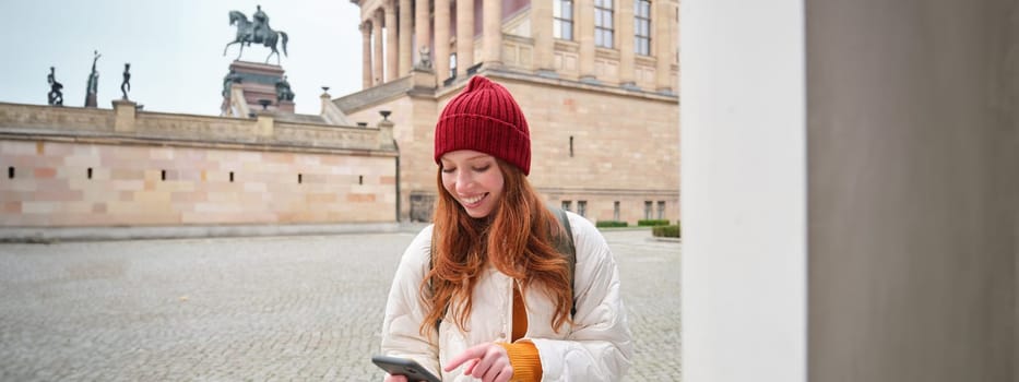 Tourism and sightseeing concept. Young redhead woman, tourist walks around city, looks at her smartphone app and at history stand, explores adventures.