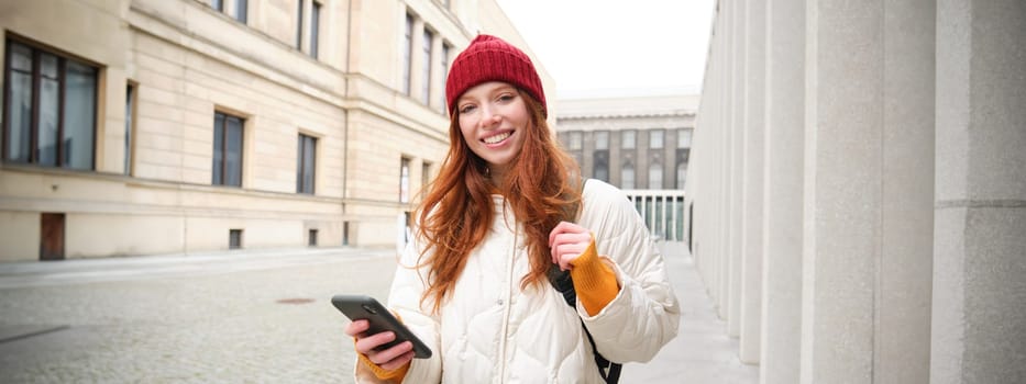 Beautiful smiling girl, tourist with backpack, holding smartphone, using map on mobile phone application, looking for sightseeing in internet app, standing outdoors.