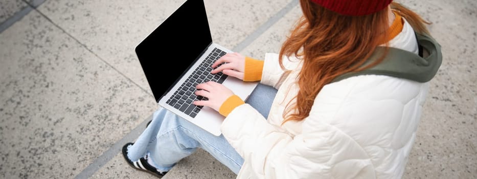Rear view of girl student typing on laptop keyboard, blank black screen for website application advertisement, sitting on stairs outdoors, connects to public wifi.