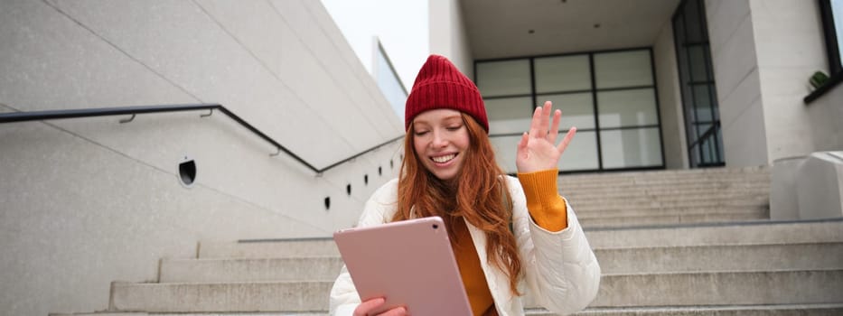 Joyful redhead teen girl, student with digital tablet, says hello, waves hand at gadget camera, connects to video chat, talks to friend in application.