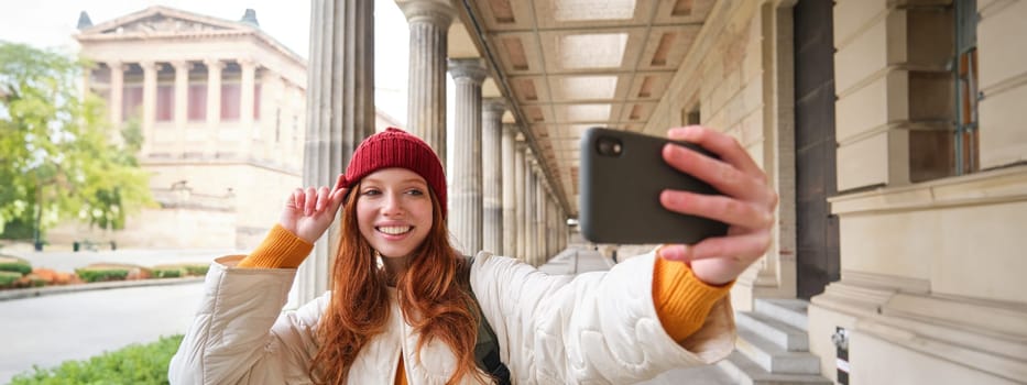Cute young redhead woman takes selfie on street with mobile phone, makes a photo of herself with smartphone app on street.