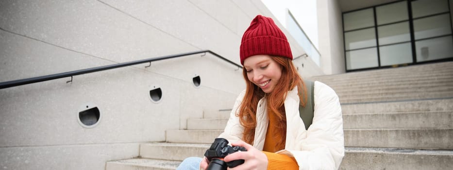 Smiling redhead girl photographer, checks her shots, holds camera and looks at screen, takes photos outdoors, walks around street and does streetstyle shooting.