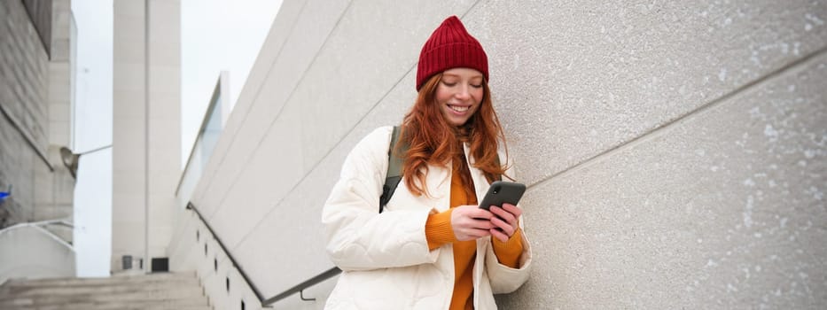 Redhead girl, young woman tourist with backpack, holds smartphone, looks for route on mobile application, searches for hotel on phone map, smiles happily.