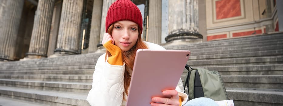 Portrait of young redhead woman sitting outdoors on stairs, reading e-book on digital tablet, wearing red hat and warm clothes.