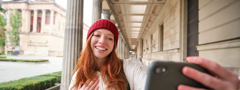 Cute young redhead woman takes selfie on street with mobile phone, makes a photo of herself with smartphone app on street.