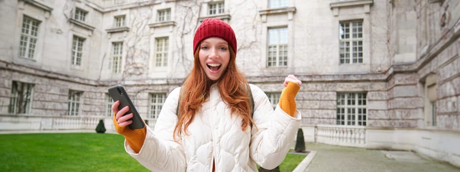 Portrait of cheerful redhead woman with smartphone, celebrating, reading amazing news and rejoicing, triumphing on street.