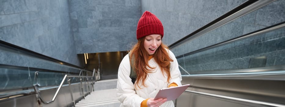 Portrait of girl student, walks up stairs from subway and looks at digital tablet with shocked, surprised face expression, reading amazing news.