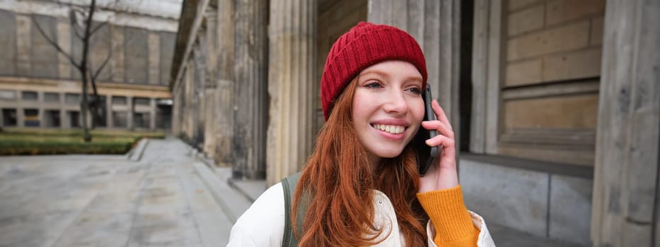 Smiling redhead female tourist talks on mobile phone and walks around city. Happy student in red hat calls friend, stands on street and uses smartphone.