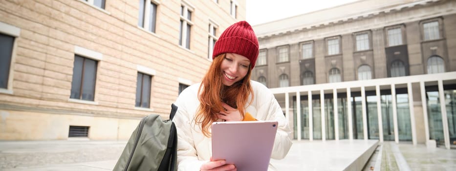 Redhead girl smiles, sits outdoors near building with digital tablet, thermos and backpack, connects to public internet and searches smth online on her gadget.