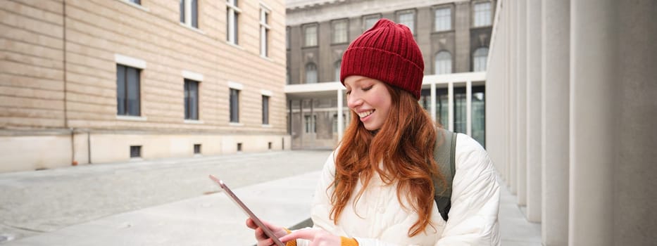 Stylish ginger girl, tourist walks with digital tablet around city, woman connects to iternet on her gadget, looking up information, texting message.