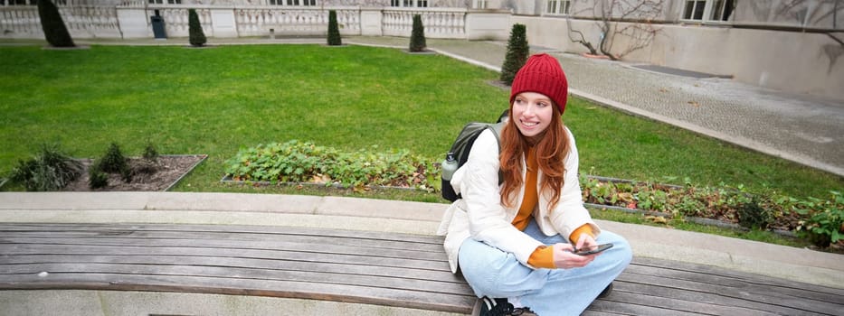 Young stylish female student with backpack, sits on bench and uses smartphone, plays on mobile phone.