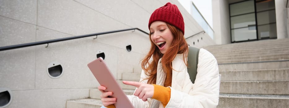 Young stylish girl, redhead female students sits on stairs outdoors with digital tablet, reads, uses social media app on gadget, plays games while waits on street.