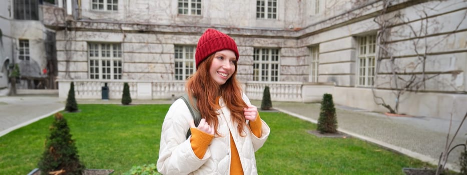 Cute ginger girl in red hat, holds backpack, walks around city and smiles. Student going to campus in University.