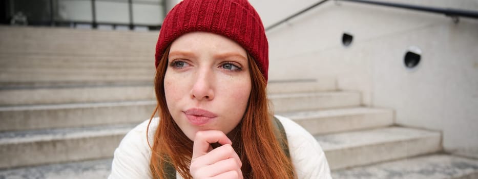Portrait of redhead girl in red hat, looks thoughtful, touches her chin, frowns and thinks with complicated face expression, sits on stairs outdoors.