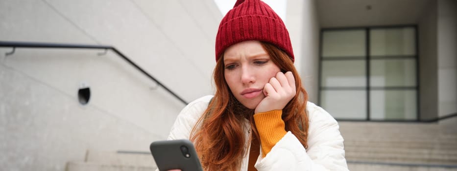 Portrait of sad redhead girl, looks upset and disappointed at smartphone screen, reads bad news text message on mobile phone and frowns, sits on stairs outdoors.