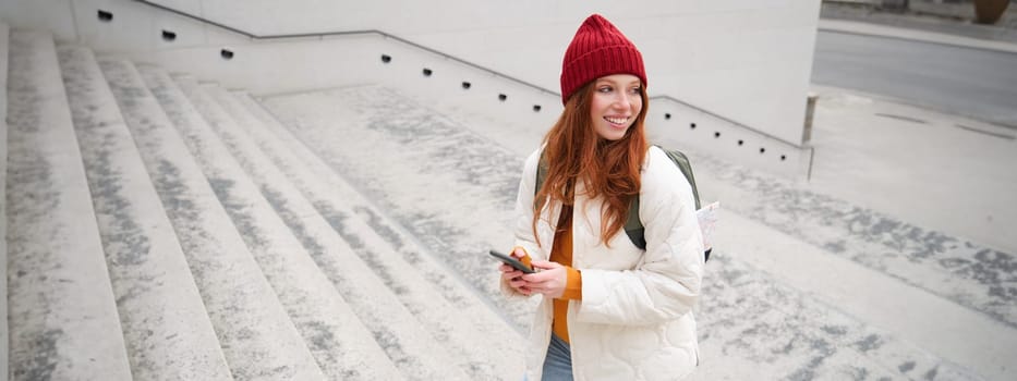 Joyful girl tourist looks at mobile phone, texts message on smartphone social media application, walks around city, looks for sightseeing on mobile app.