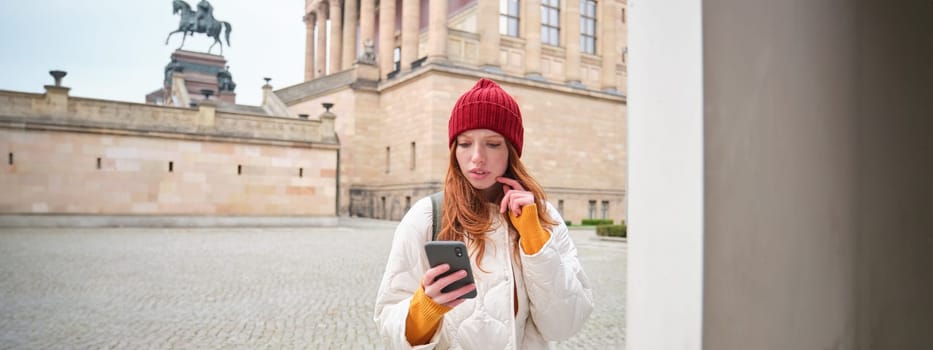 Smiling redhead girl tourist, walks around city and explores popular landmarks, sightseeing, holding smartphone, looking at her mobile app and checks with city map.