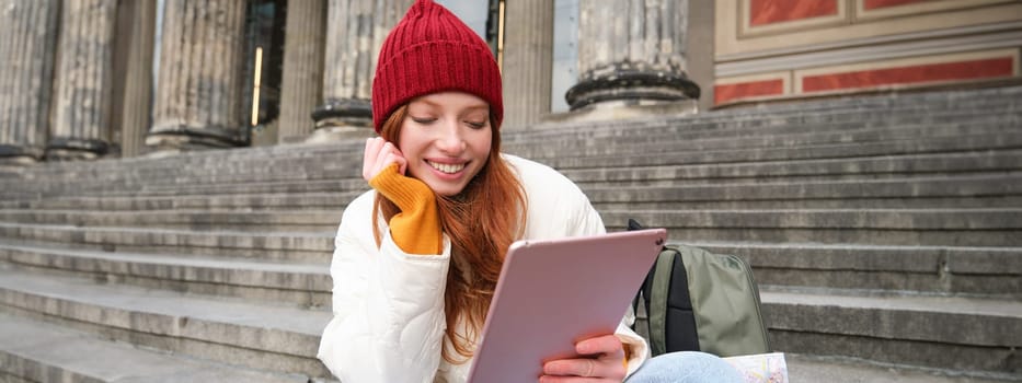 Beautiful young modern girl with red hair, holds digital tablet, sits on stairs near museum and connects public internet, sends message on gadget app.