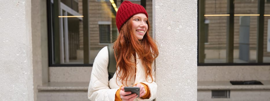 Beautiful redhead girl with smartphone, standing on street, holding mobile phone, using taxi app, connects to public wifi and uses internet, sends a text message.