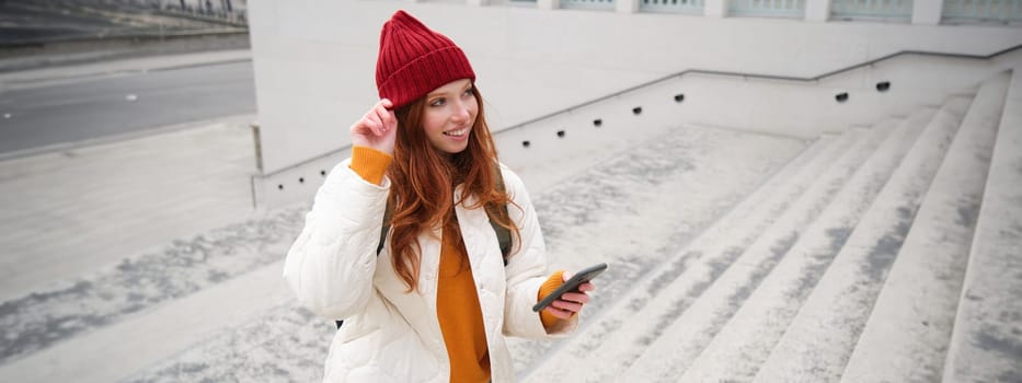 Joyful girl tourist looks at mobile phone, texts message on smartphone social media application, walks around city, looks for sightseeing on mobile app.
