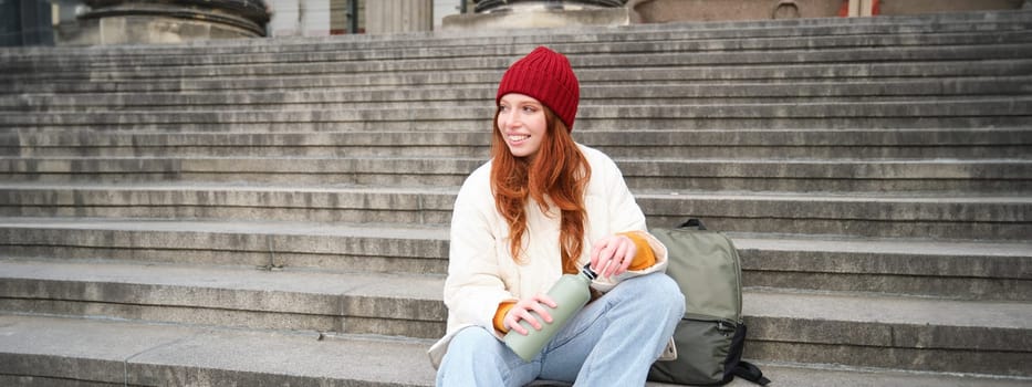 Young redhead female tourist rests during her trip, opens thermos and drinks hot tea, having a break after sightseeing.
