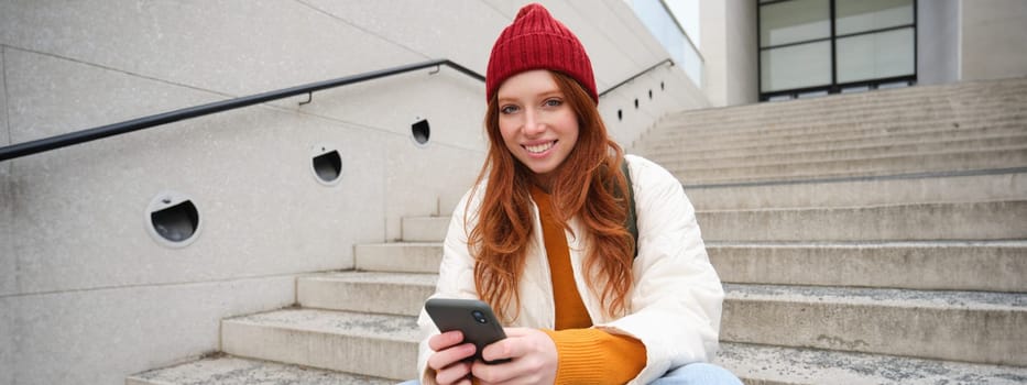 Beautiful redhead teen girl, sits on stairs and smiles, holds mobile phone, uses smartphone application, chats on telephone app.