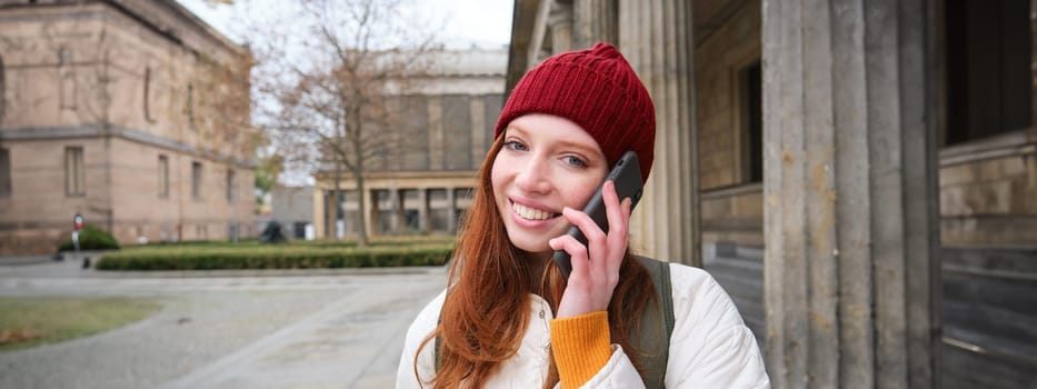 Smiling young redhead woman listens to voice message, makes a phone call, walks on street and talks to someone on smartphone.
