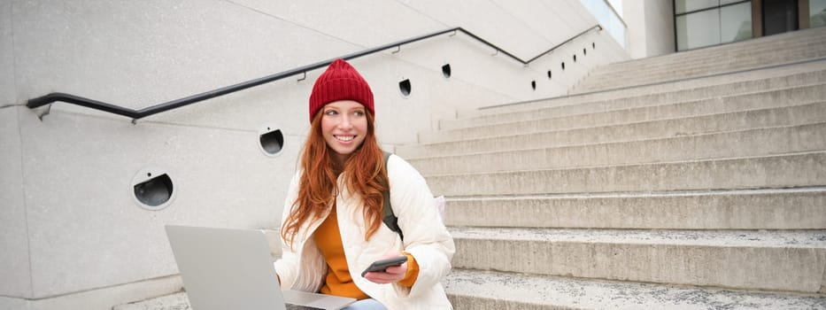 Smiling redhead woman with mobile phone and laptop, sitting on stairs outside building, connects to public wifi, using smartphone and computer.