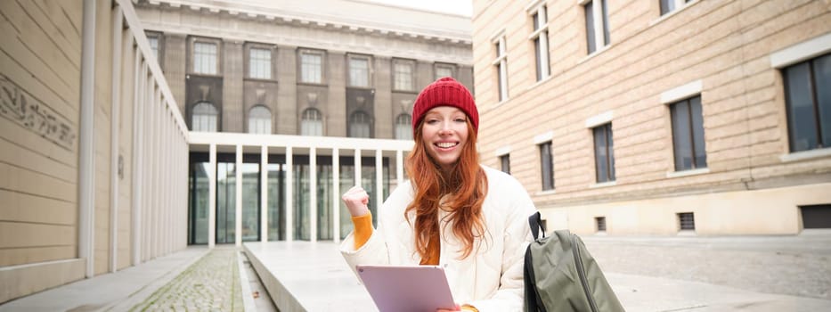 Beautiful redhead woman in red hat, sits with backpack and thermos, using digital tablet outdoors, connects to wifi, texts message, books tickets online.
