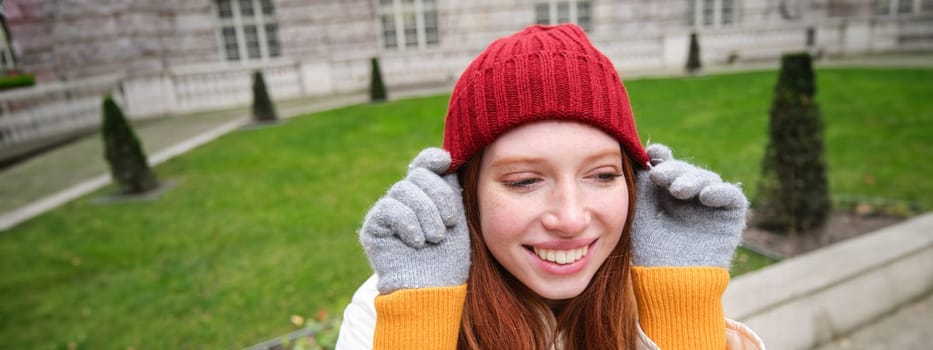 Close up portrait of beautiful redhead woman in red knitted hat, warm gloves, smiling and looking happy at camera, sitting in park.