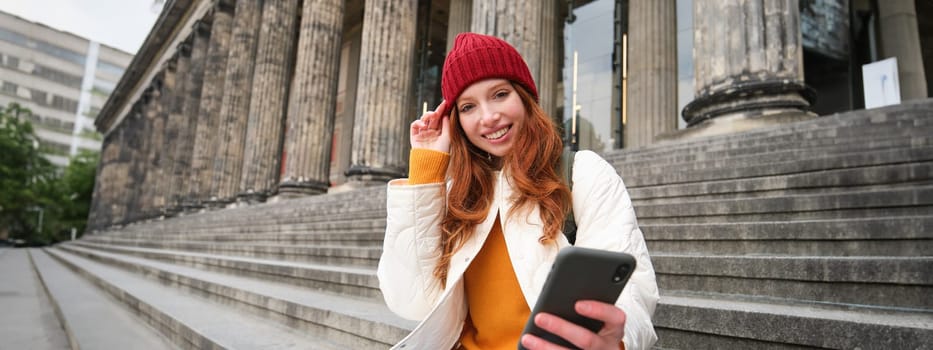 Portrait of young redhead woman in red hat, sitting on stairs, tourist looks at her mobile phone, rests on staircase of museum, connects public wifi.