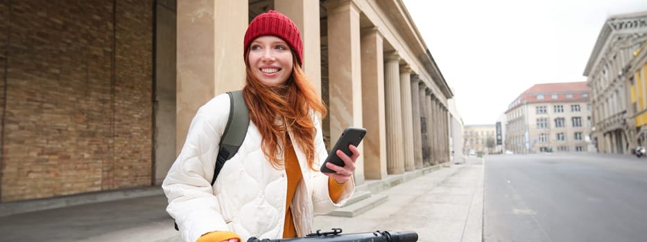 Redhead girl, tourist with backpack, uses mobile phone to rent e-scooter on streets of European city.