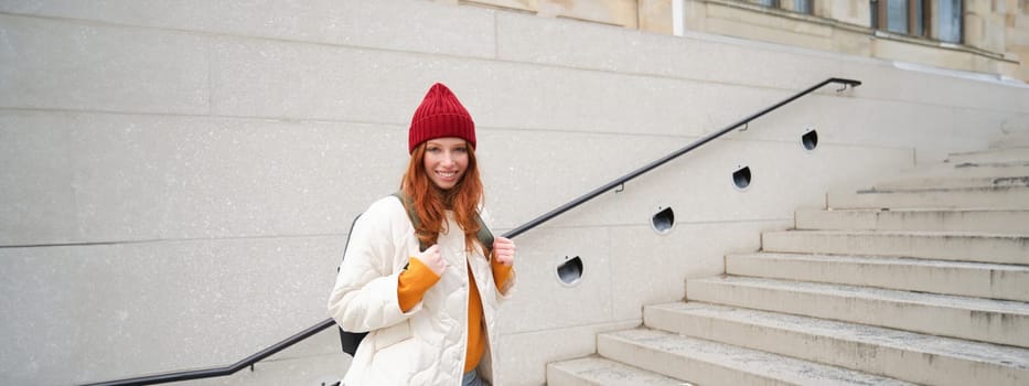 Happy young girl, redhead tourist in red hat with backpack, walks around town, explores city, backpacking around europe, travelling alone.