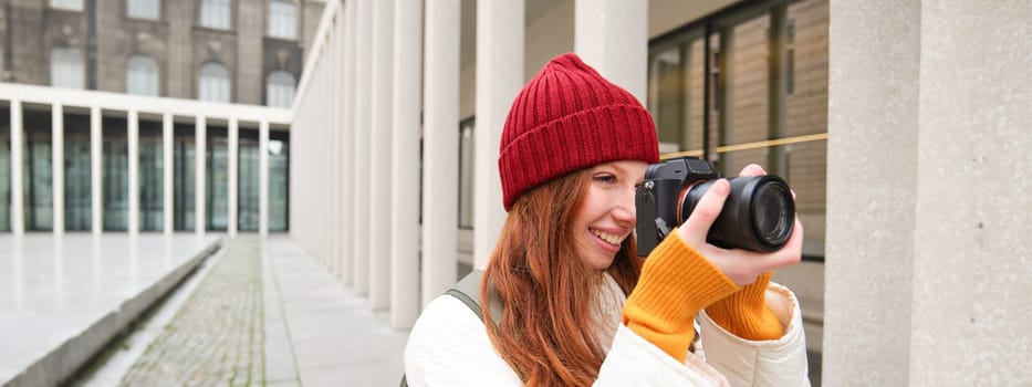Smiling redhead girl photographer, taking pictures in city, makes photos outdoors on professional camera. Young talent and hobby concept