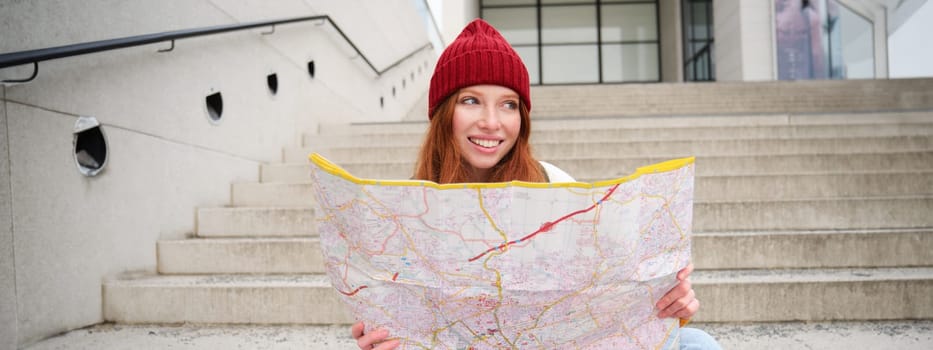 Young smiling redhead girl, tourist sits on stairs outdoors with city paper map, looking for direction, traveller backpacker explores city and looks for sightseeing.