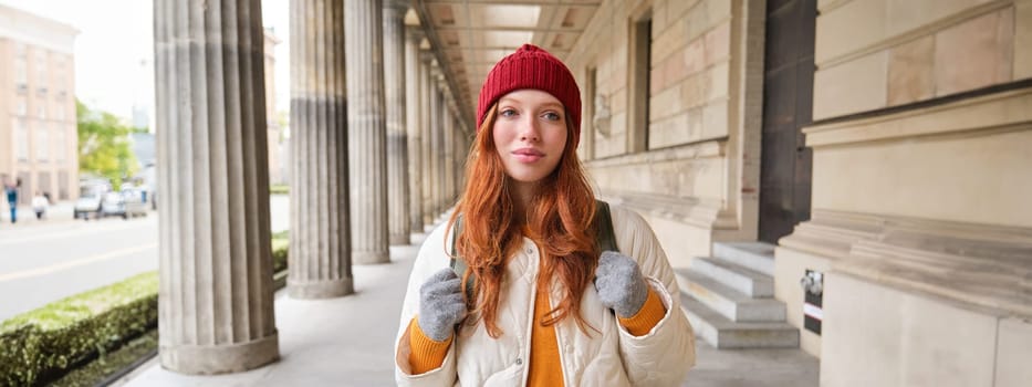 Smiling redhead girl with backpack, walks in city and does sightseeing, explore popular landmarks on her tourist journey around Europe.