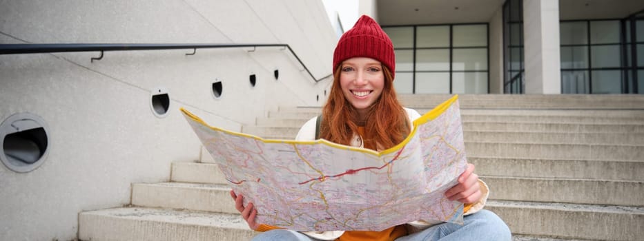 Beautiful girl tourist sits on stairs with city map, plans her journey, looks for direction while travelling around town, searches route for sightseeing.