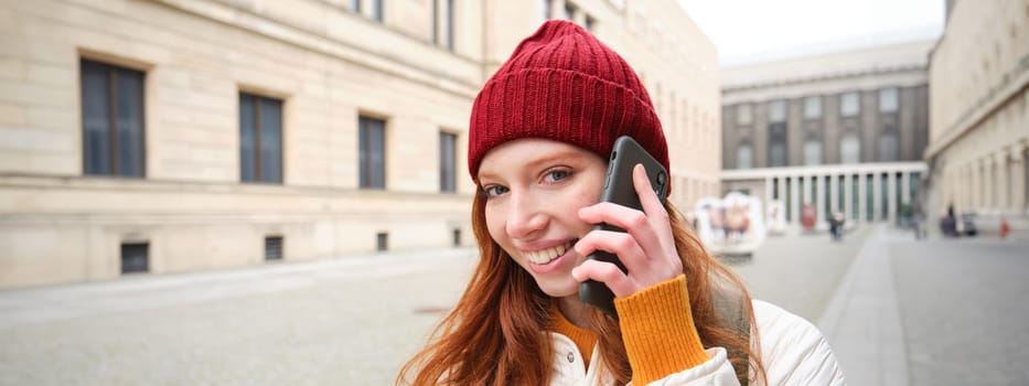 Mobile broadband and people. Smiling young redhead woman walks in town and talks on mobile phone, calling friend on smartphone, using internet to make a call abroad.