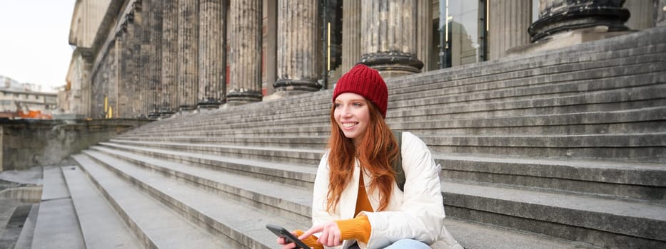 Stylish young redhead woman, talking on mobile phone app, using social media application, looking for something online on smartphone, sits on stairs outdoors.